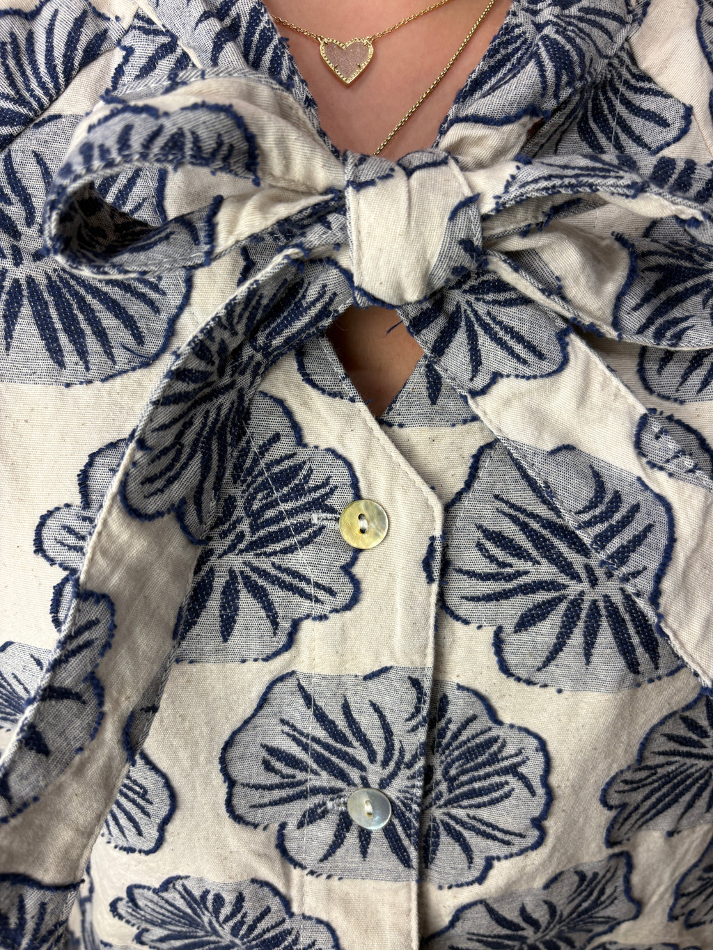 Close-up of a person wearing the Dawn Blouse in cream-colored linen shirt with a navy blue floral block print pattern, featuring a large bow at the collar and a button-down front.