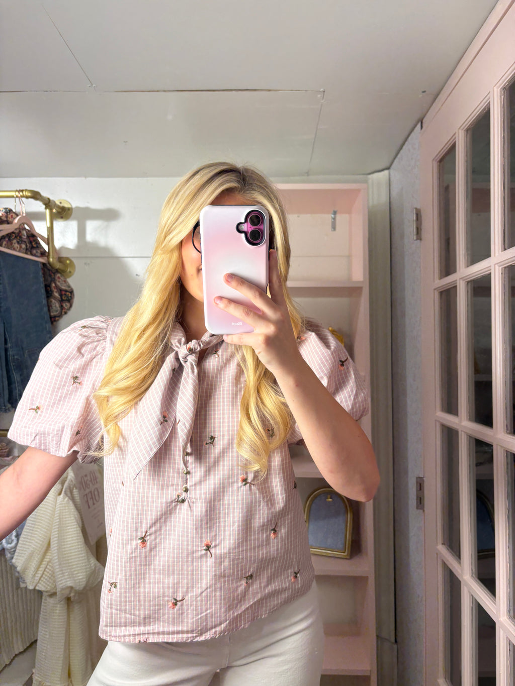 A woman takes a mirror selfie wearing a light pink and white gingham short-sleeve blouse with a tie-neck and small embroidered details, paired with white pants in a pink-toned room.