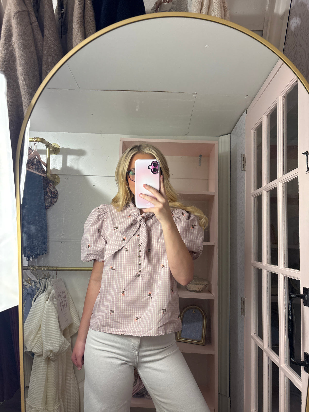A woman takes a mirror selfie in a fitting room wearing a light pink and white gingham short-sleeve blouse with a bow tie neckline and small embroidered details, paired with white pants.