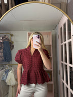 A woman takes a mirror selfie in a boutique showing a short-sleeve burgundy eyelet lace peplum blouse and white pants.
