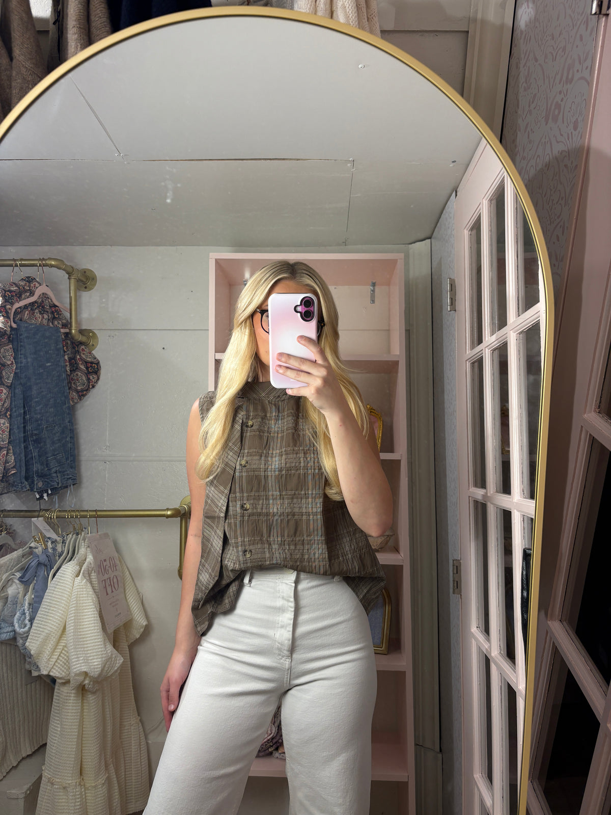 A woman taking a mirror selfie in a room, wearing a brown and beige plaid sleeveless top and white pants.