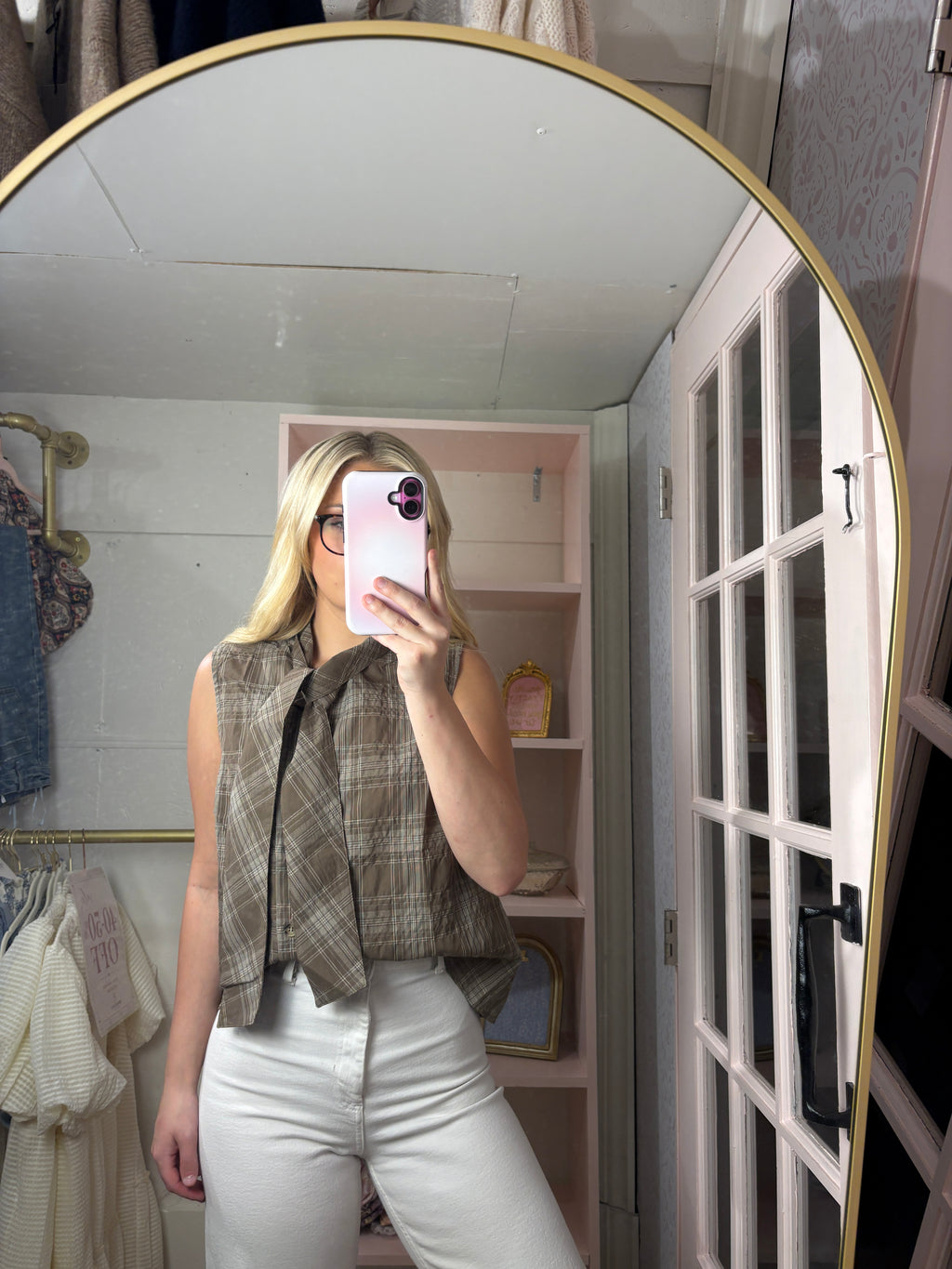A woman takes a mirror selfie in a dressing room wearing a brown and beige plaid sleeveless top with a large bow detail and white pants.