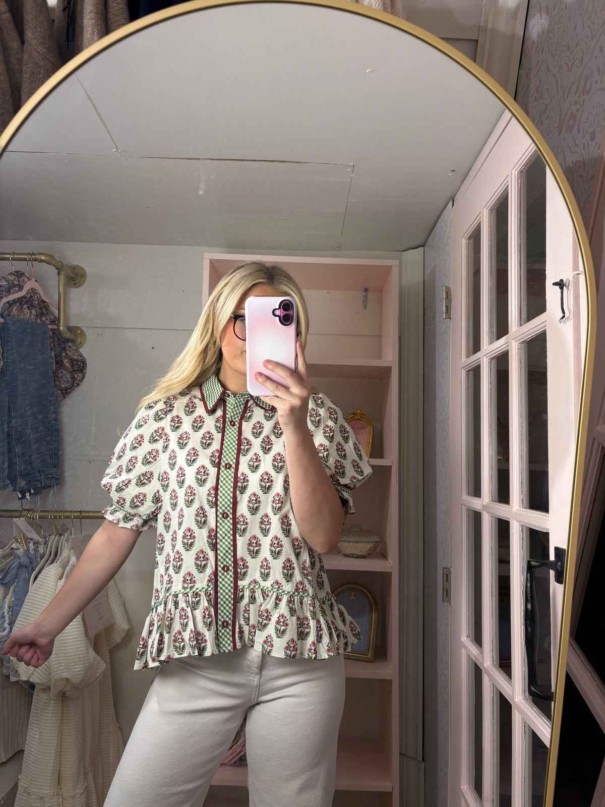 A woman takes a mirror selfie in a fitting room wearing a short-sleeve button-up blouse with a green, red, and white floral pattern, paired with light beige pants.