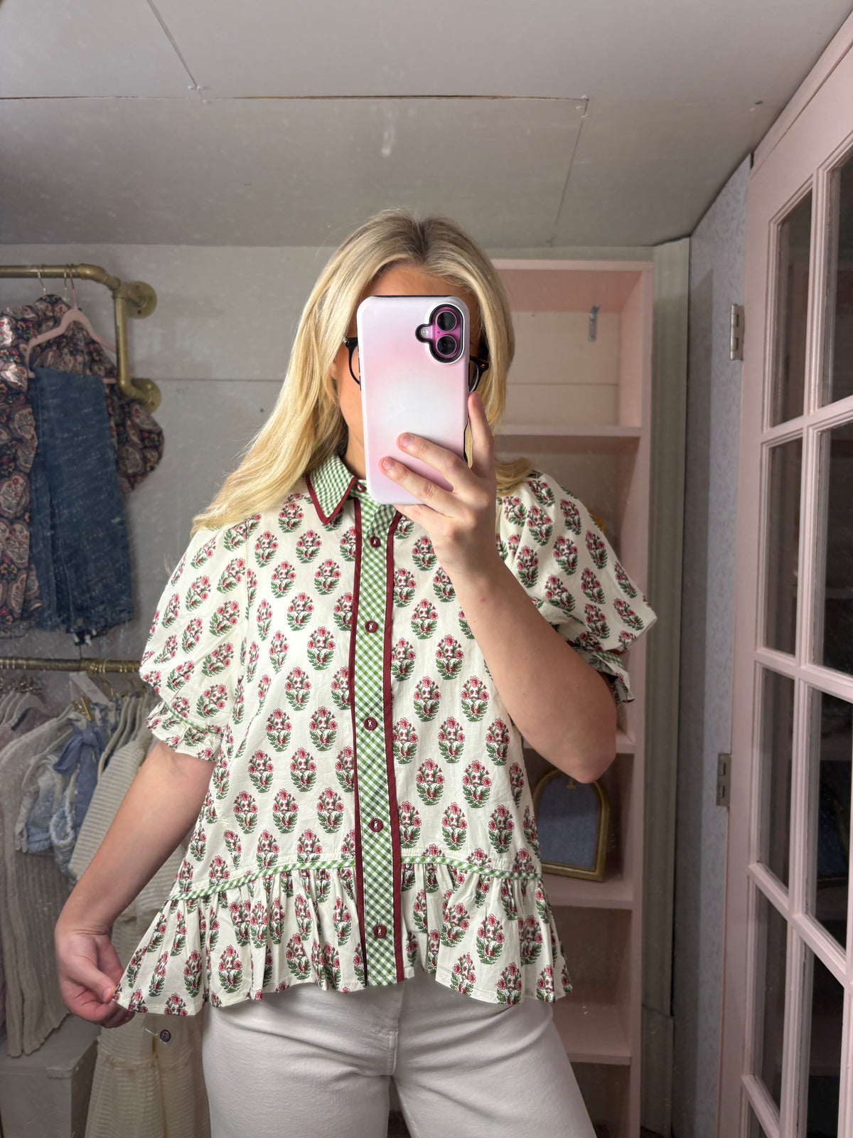 A woman takes a mirror selfie wearing a short-sleeve button-up blouse with a green and red floral print on a cream background, paired with light beige pants.