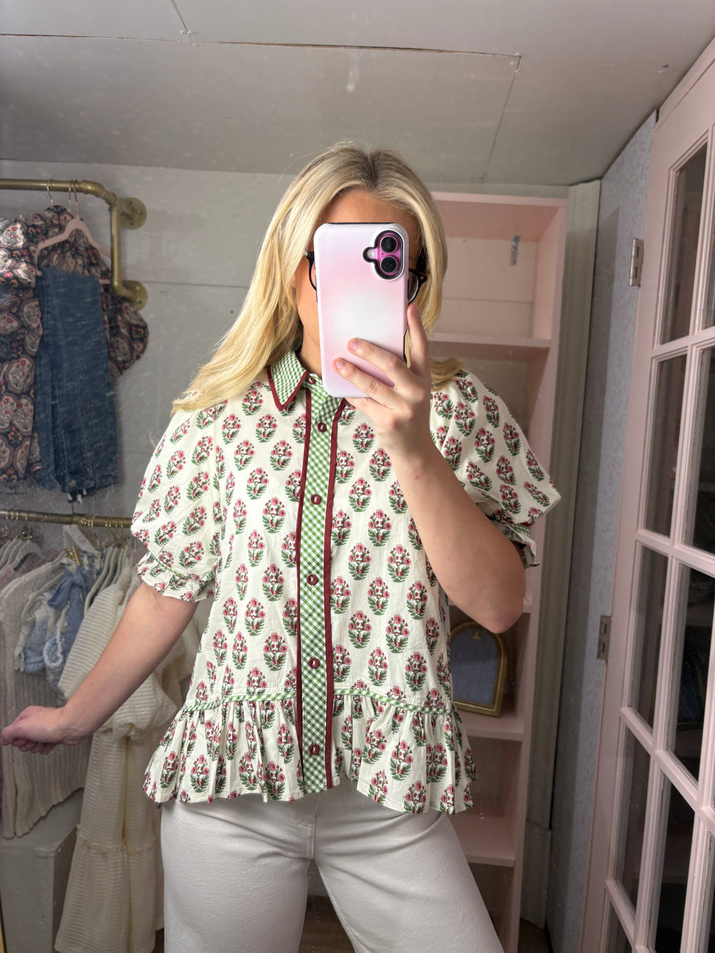 A woman takes a mirror selfie in a fitting room wearing a cream-colored short-sleeve blouse with a green and red floral print, a green patterned tie, and white pants.