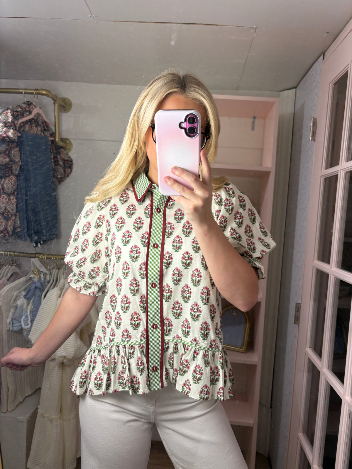 A woman takes a mirror selfie in a fitting room wearing a cream-colored short-sleeve blouse with a green and red floral print, a green patterned tie, and white pants.