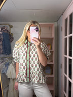 A woman takes a mirror selfie wearing a short-sleeve button-up blouse with a green and red floral pattern on a cream background, paired with light gray pants.
