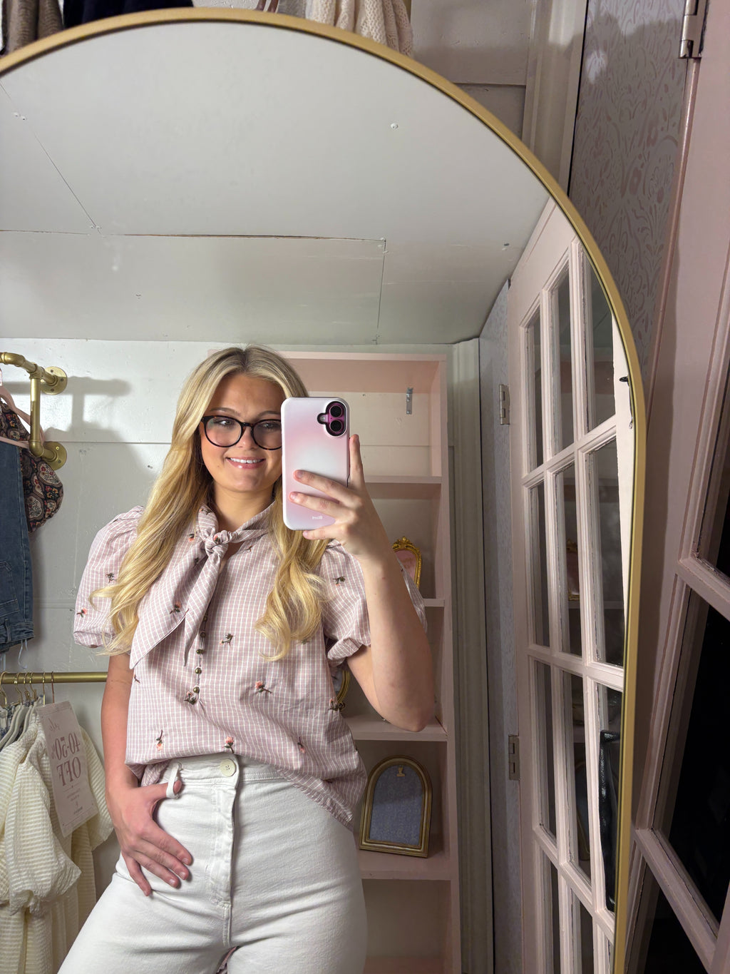 A woman takes a mirror selfie in a dressing room, wearing a light pink and white gingham short-sleeve blouse with a bow tie neckline and white pants.