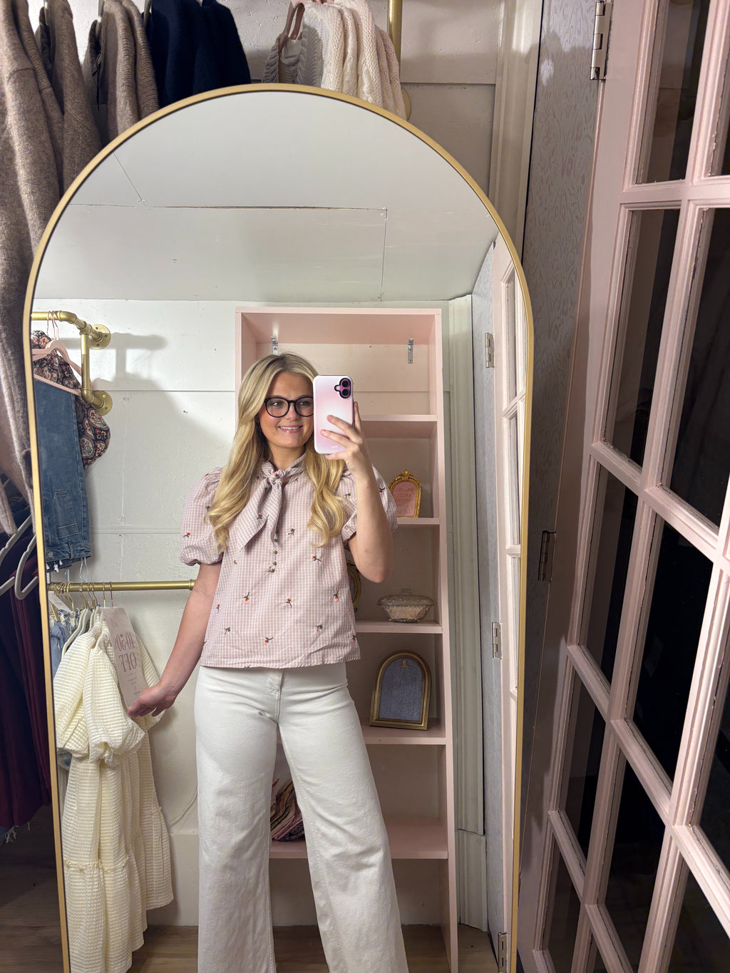 A woman takes a mirror selfie in a dressing room wearing a light pink short-sleeve blouse with a bow tie neckline and small embroidered details, paired with white wide-leg pants. The room features a large arched gold-framed mirror, a pink shelving unit, and hanging garments.