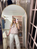 A woman takes a mirror selfie in a dressing room wearing a light pink short-sleeve blouse with a bow tie neckline and small embroidered details, paired with white wide-leg pants. The room features a large arched gold-framed mirror, a pink shelving unit, and hanging garments.