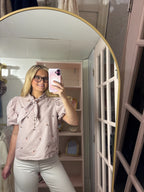 A woman takes a mirror selfie in a pink and white gingham short-sleeve blouse with a bow tie collar and small embroidered details, paired with white pants in a pink dressing room.