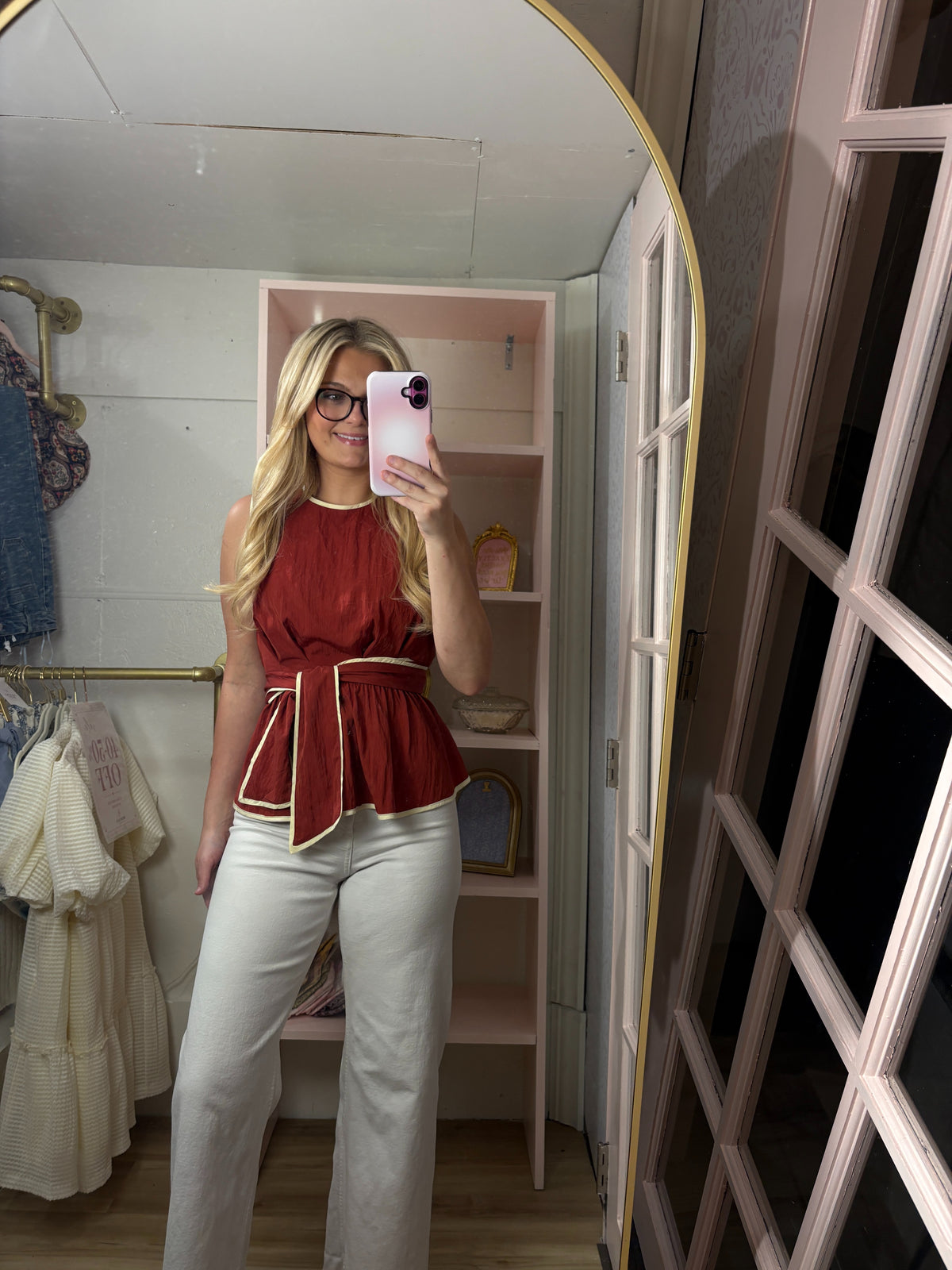 A woman takes a mirror selfie in a fitting room wearing a sleeveless red peplum top with cream piping and white pants.
