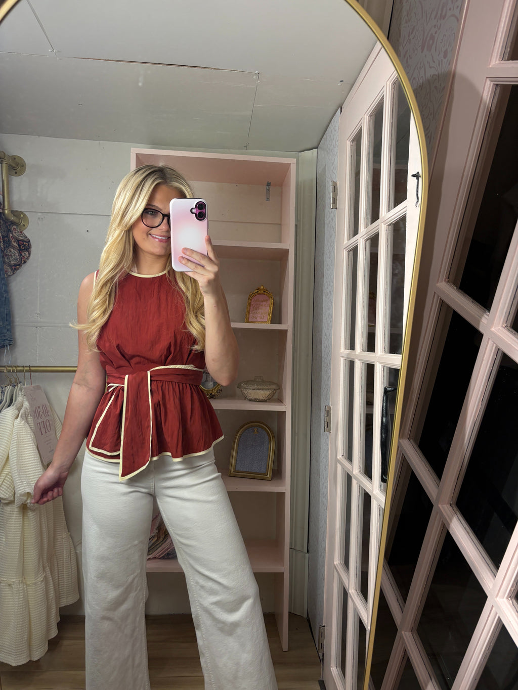 A woman takes a mirror selfie in a boutique fitting room, wearing a rust red sleeveless peplum top with gold piping and white wide-leg pants.