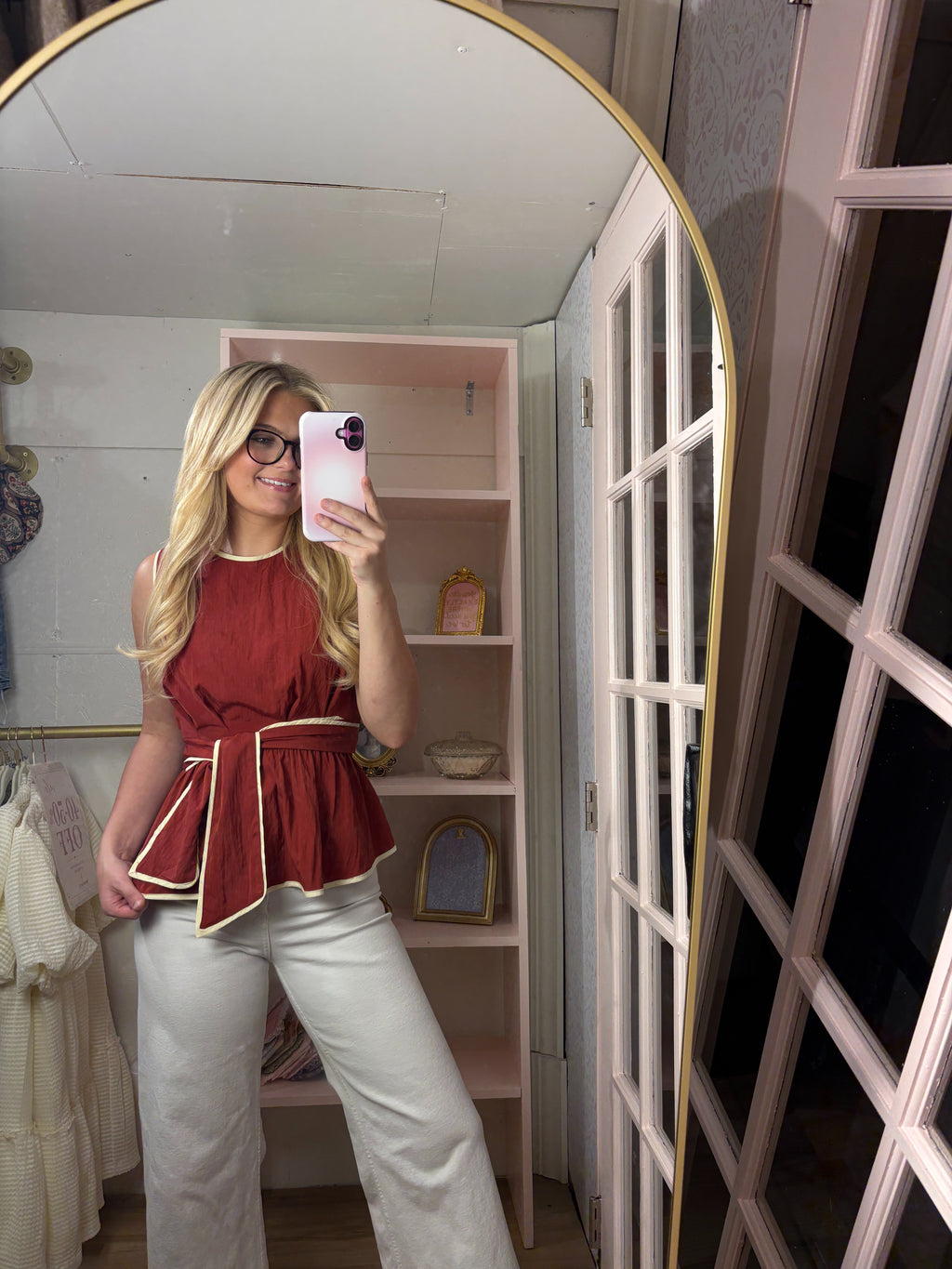 A woman takes a mirror selfie in a dressing room wearing a sleeveless rust-red peplum top with cream trim and white wide-leg pants.