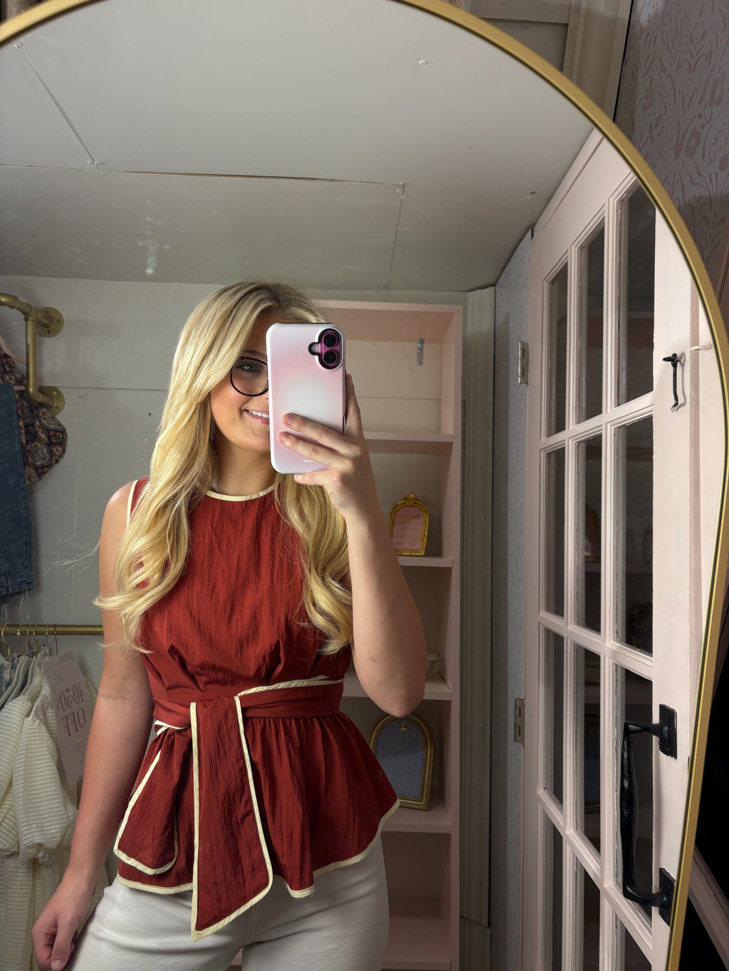 A woman takes a mirror selfie in a dressing room wearing a sleeveless rust-red peplum top with cream piping and a matching waist tie, paired with white pants.