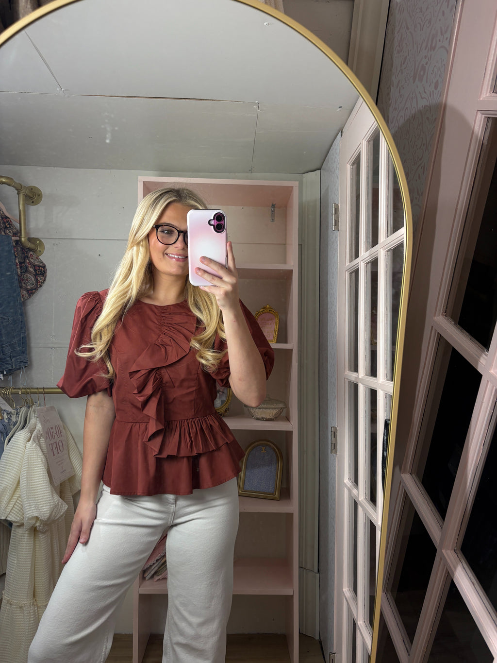 A woman takes a mirror selfie in a room with pink shelving, wearing a rust-brown ruffled short-sleeve blouse and white pants.