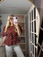 A woman takes a mirror selfie in a fitting room wearing a rust-brown ruffled short-sleeve peplum blouse and white pants.
