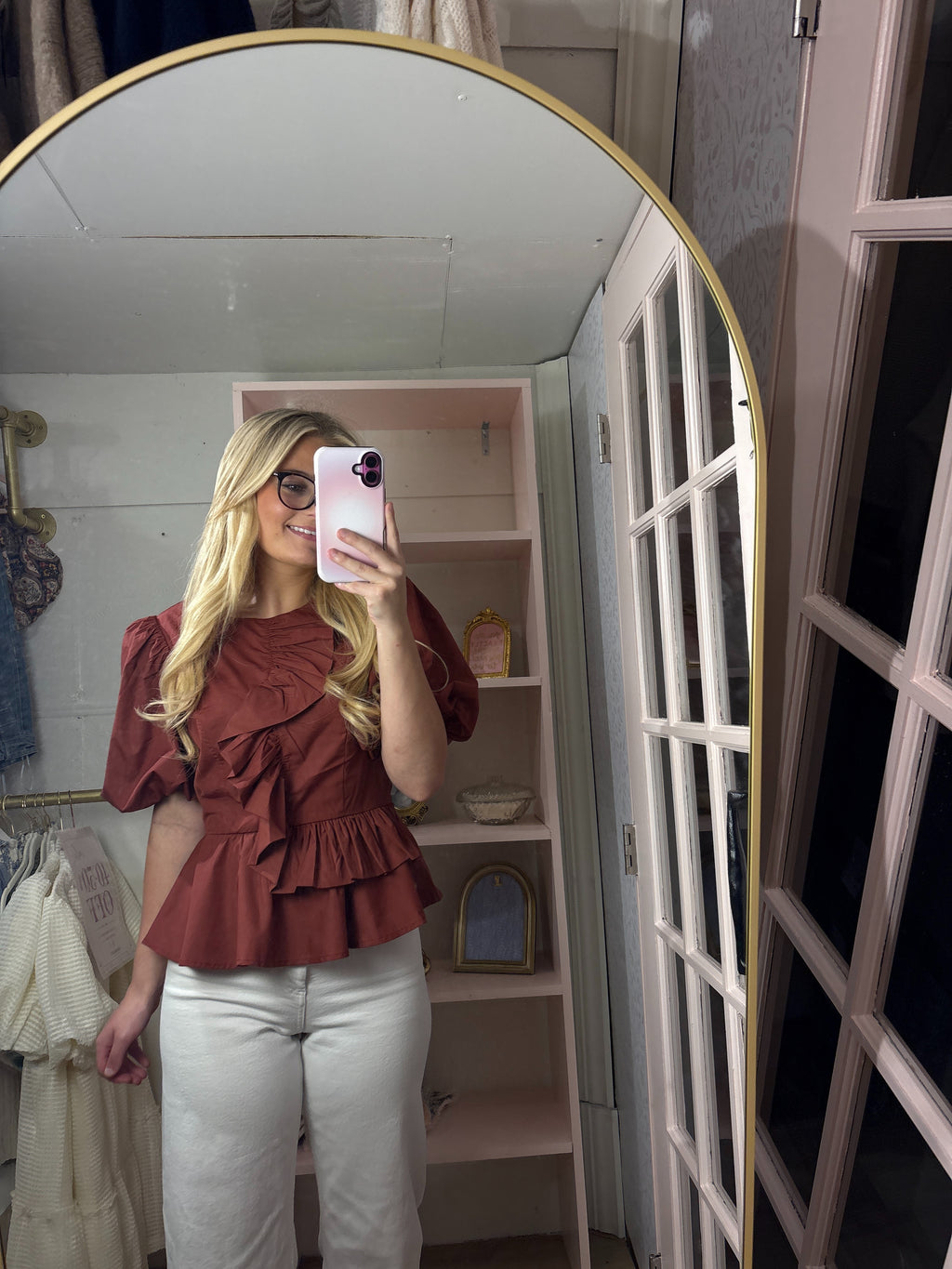 A woman taking a mirror selfie in a fitting room, wearing a rust-brown ruffled short-sleeve blouse and white pants.