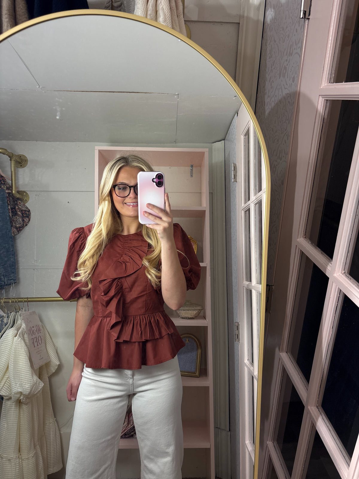 A woman takes a mirror selfie in a boutique, wearing a rust-brown ruffled short-sleeve blouse and white pants. The background shows a pink shelving unit and a large gold-framed mirror.