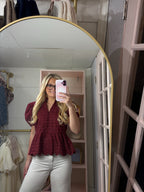 A woman takes a mirror selfie in a pink dressing room, wearing a burgundy eyelet lace peplum blouse and white pants.