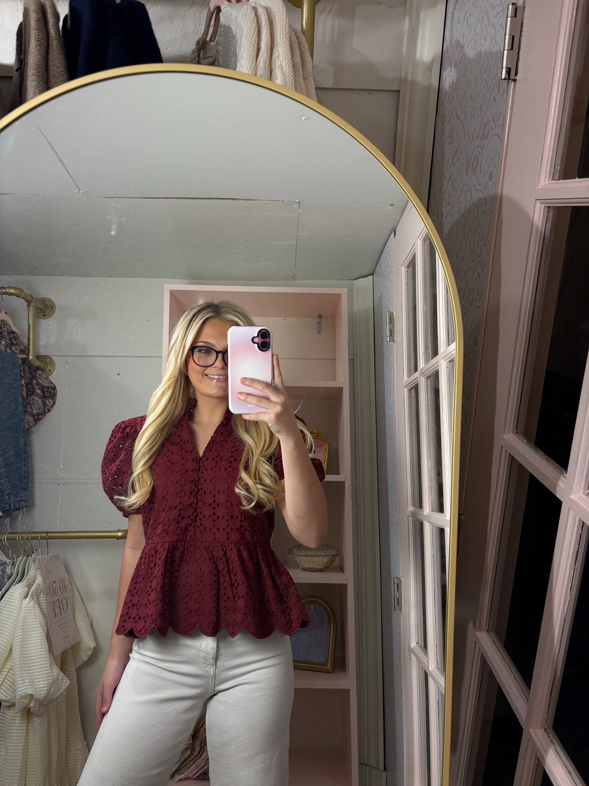 A woman takes a mirror selfie in a pink dressing room, wearing a burgundy eyelet lace peplum blouse and white pants.
