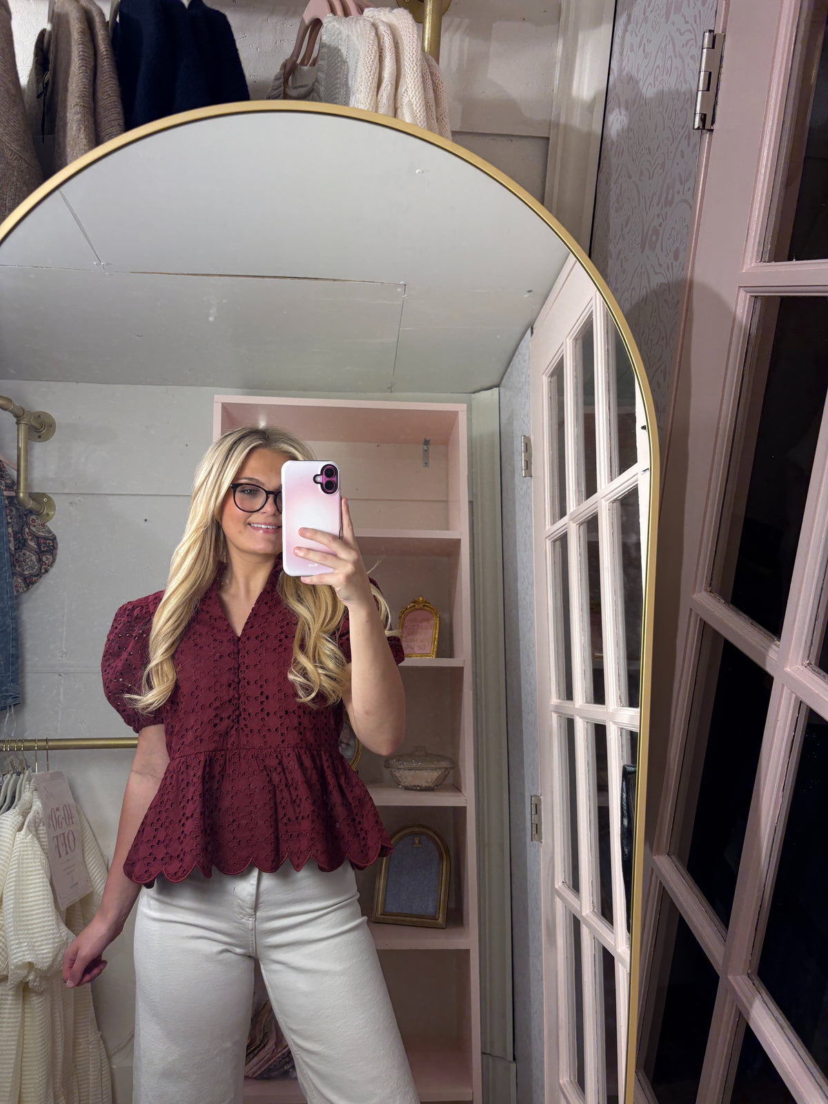 A woman takes a mirror selfie in a fitting room wearing a burgundy eyelet puff-sleeve peplum top and white pants.