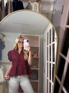 A woman takes a mirror selfie in a dressing room wearing a burgundy eyelet lace peplum blouse and white pants.