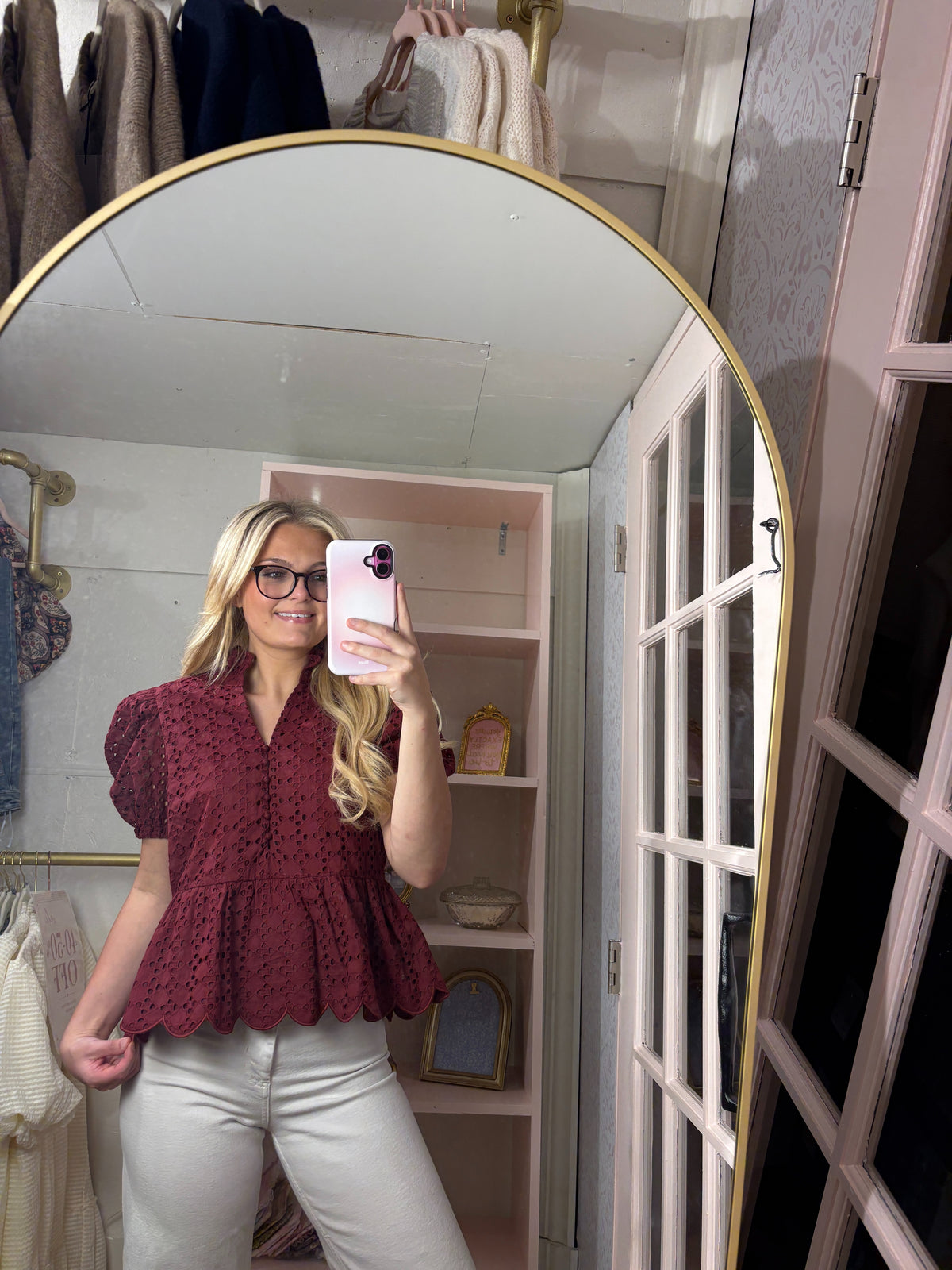 A woman takes a mirror selfie in a dressing room wearing a burgundy eyelet lace peplum blouse and white pants.