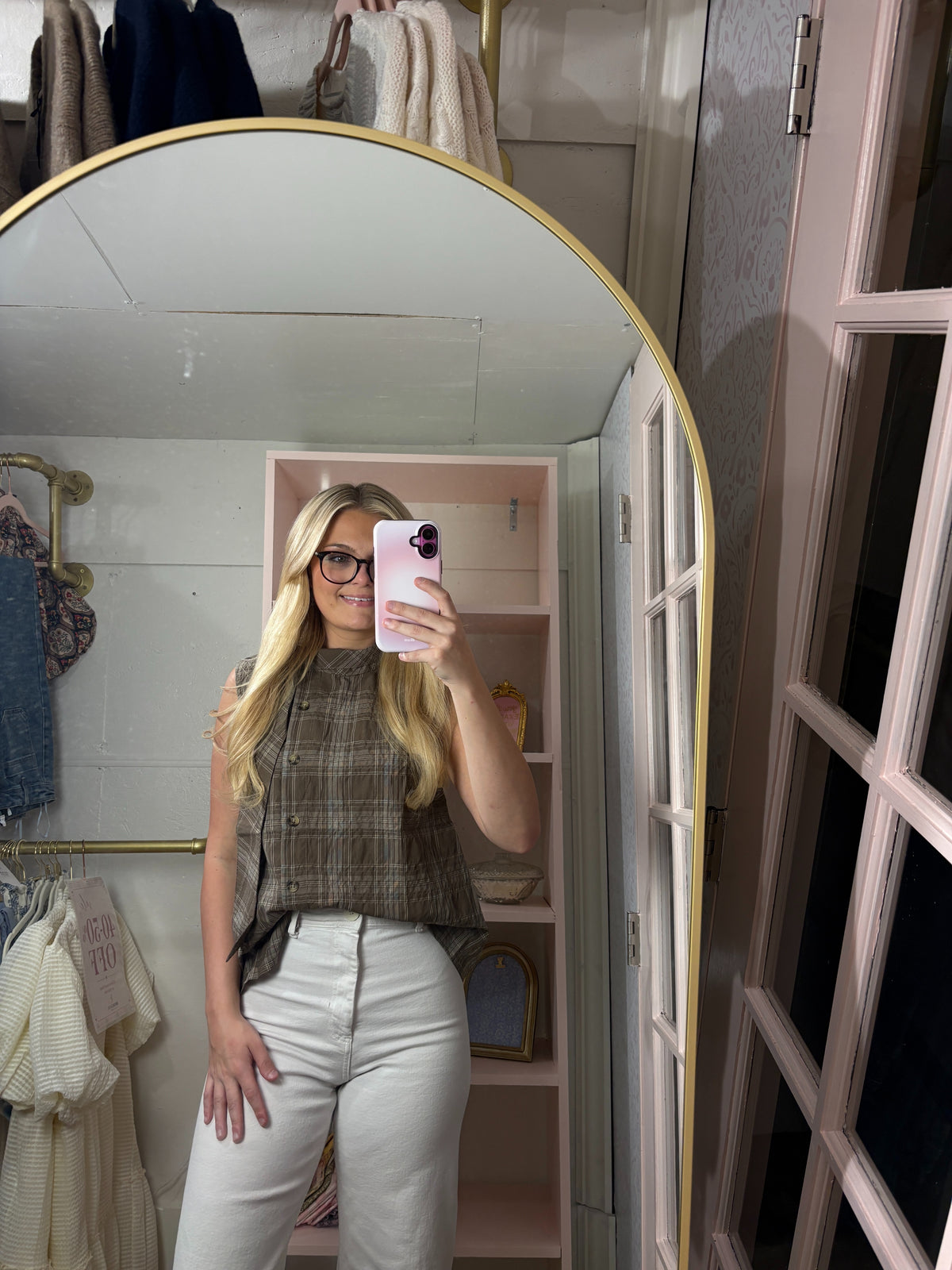 A woman takes a mirror selfie in a fitting room wearing a sleeveless brown and gray plaid top with white pants. The background shows a pink shelving unit and clothing racks.