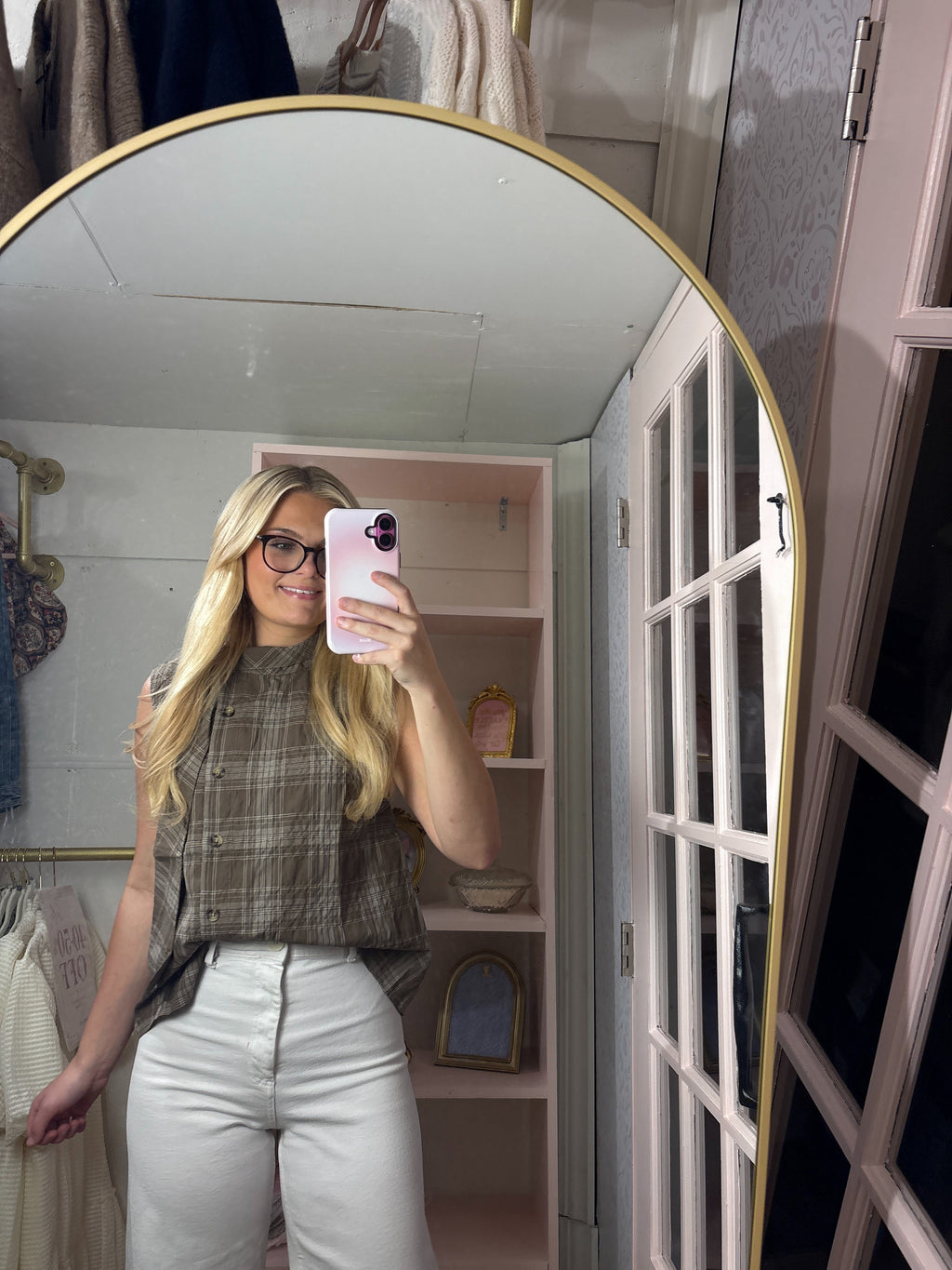 A woman takes a mirror selfie in a dressing room, wearing a brown and gray plaid sleeveless top and white pants. The background features a large arched gold-framed mirror, a pink shelving unit, and a white French door.