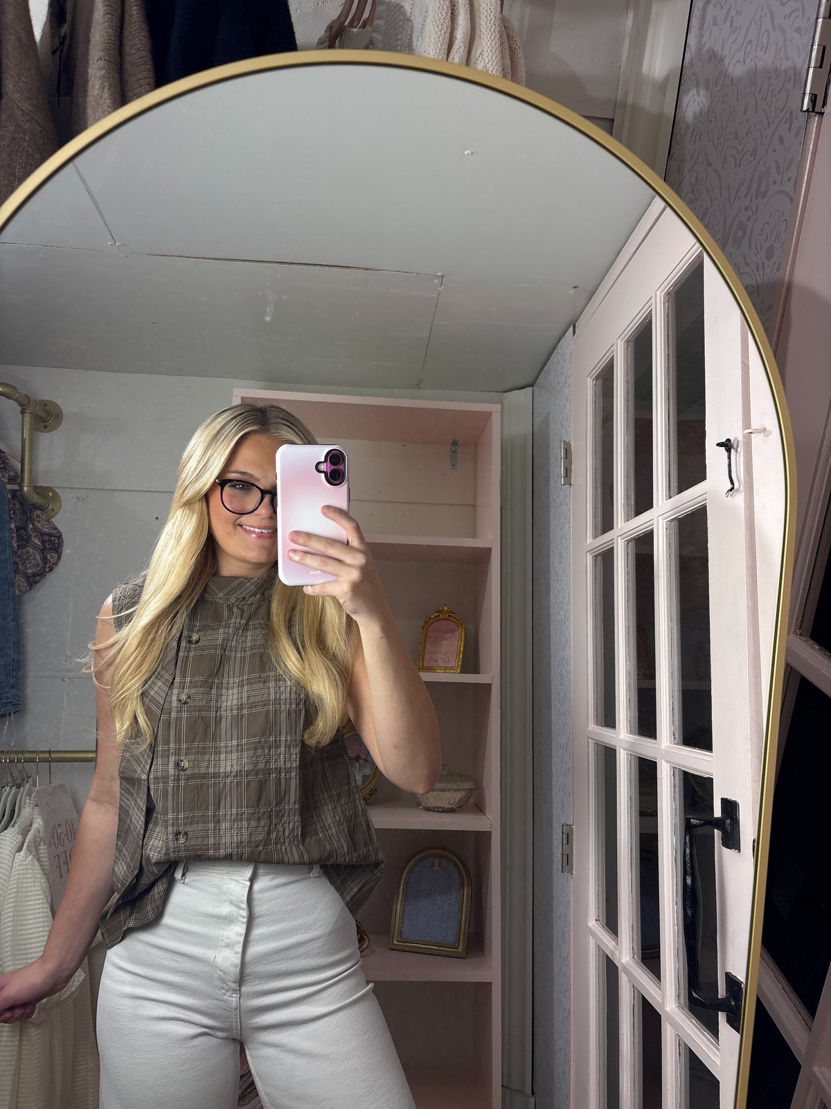 A woman takes a mirror selfie in a dressing room, wearing a sleeveless brown and beige plaid button-up top and white pants. The image is framed by a large arched gold-framed mirror, with shelves and clothing visible in the background.