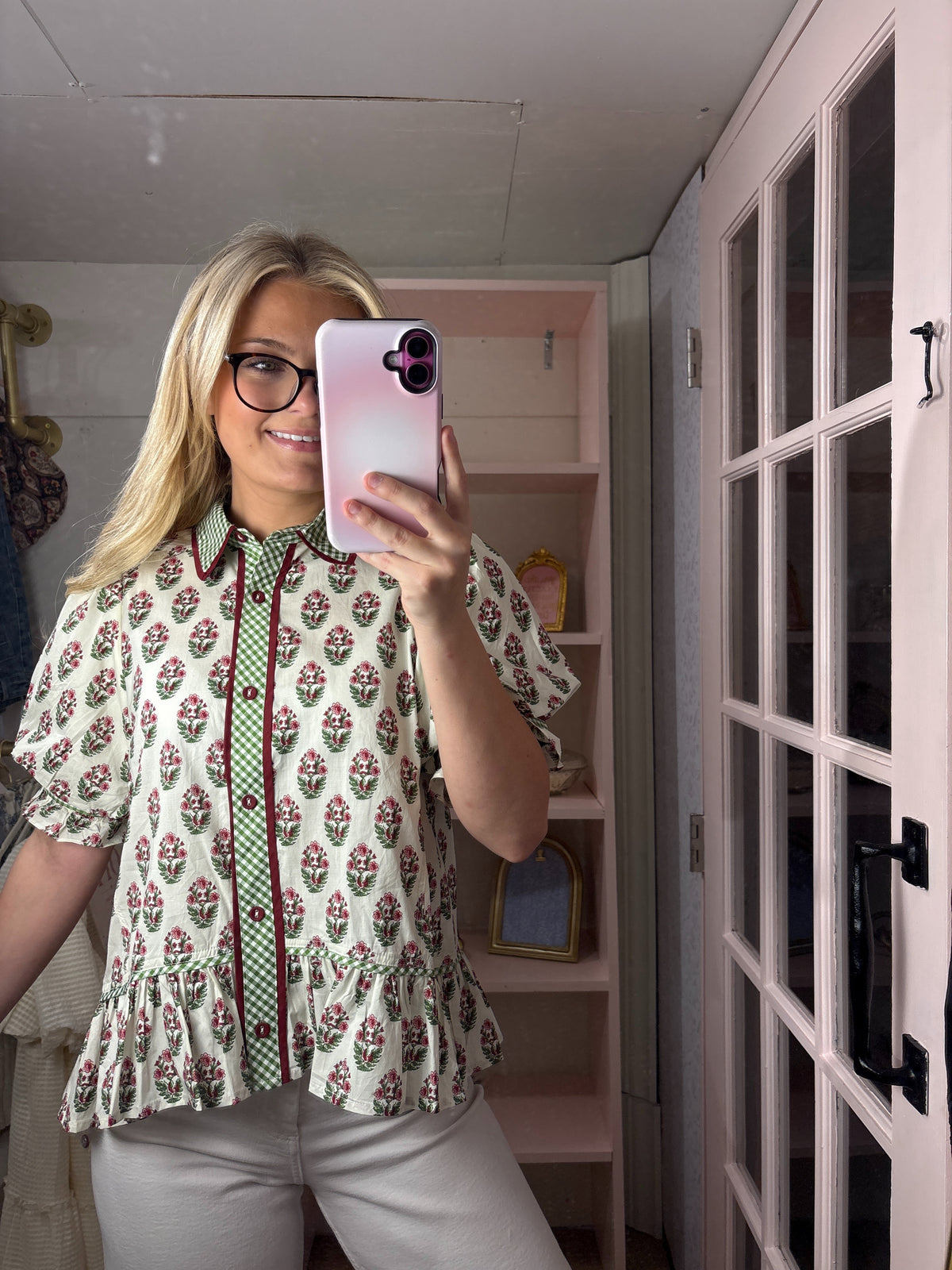 A woman taking a mirror selfie in a room, wearing a short-sleeve button-up blouse with a green, red, and white floral pattern and ruffled hem, paired with light gray pants.
