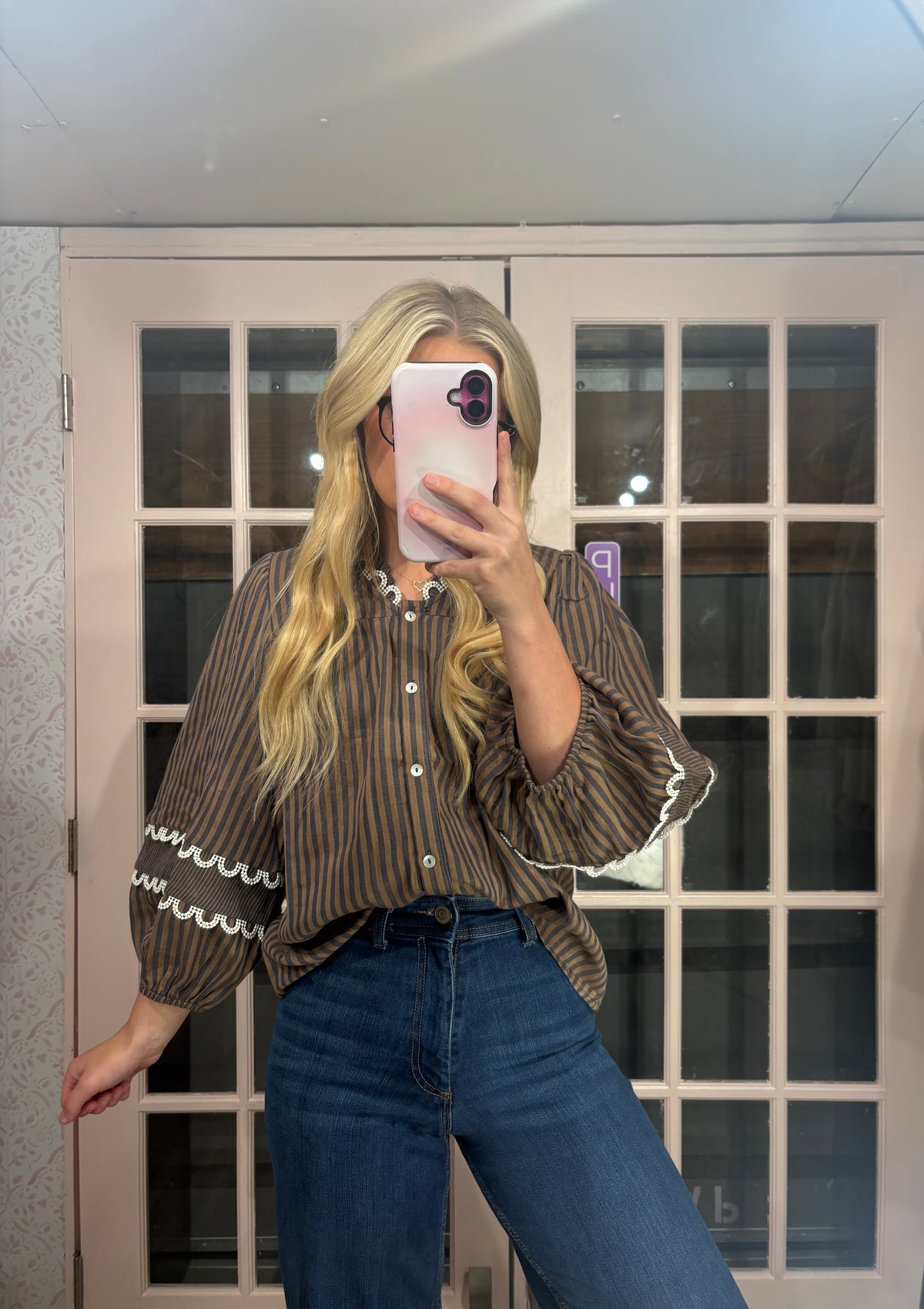 A woman takes a mirror selfie wearing a brown and beige vertically striped blouse with white ruffle trim on the collar and cuffs, paired with blue jeans.