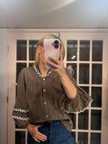 A woman takes a mirror selfie wearing a brown and black vertically striped blouse with white scalloped trim on the collar and cuffs, paired with blue jeans.
