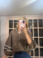 A woman takes a mirror selfie wearing a brown and black vertically striped blouse with white ruffle trim on the collar and cuffs, paired with blue jeans.