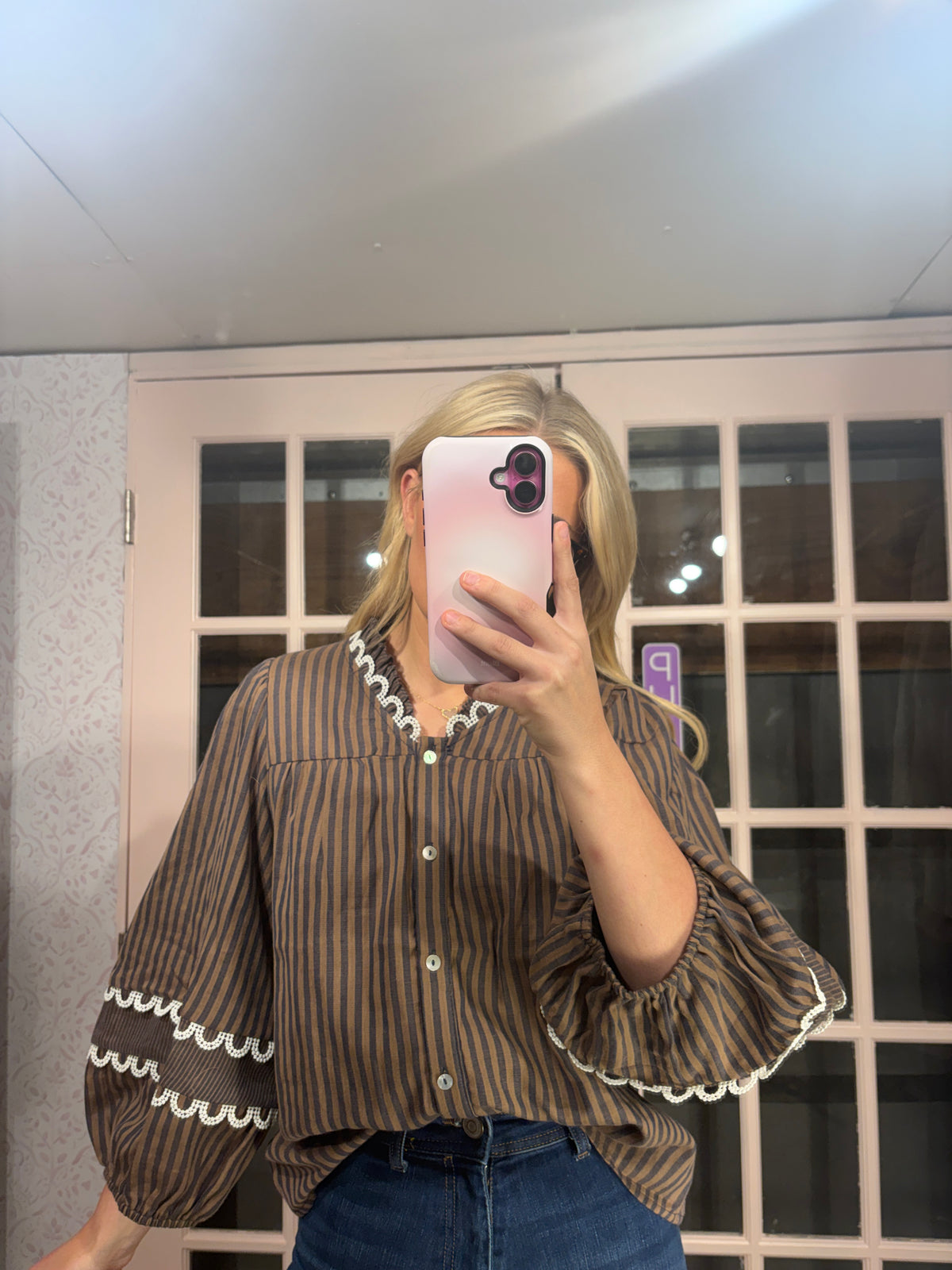 A woman takes a mirror selfie wearing a brown and black vertically striped blouse with white ruffle trim on the collar and cuffs, paired with blue jeans.