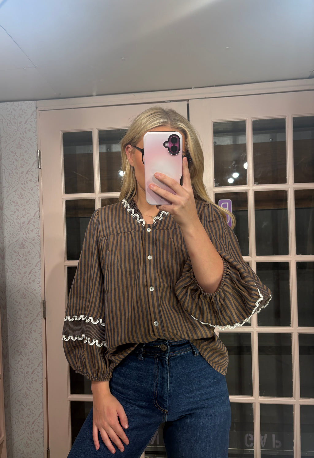A woman takes a mirror selfie wearing a brown and black vertically striped blouse with white ruffle trim on the collar and cuffs, paired with blue jeans.