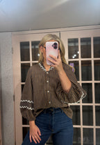 A woman takes a mirror selfie wearing a brown and black vertically striped blouse with white ruffle trim on the collar and cuffs, paired with blue jeans.