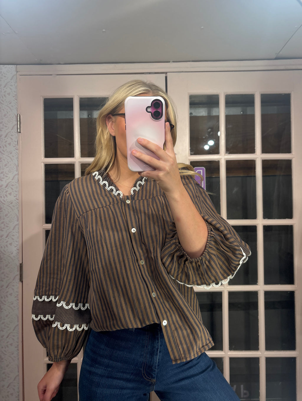 A woman takes a mirror selfie wearing a brown and black vertically striped blouse with white scalloped trim on the collar and cuffs, paired with blue jeans.