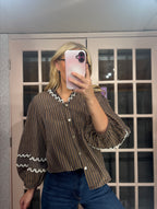A woman takes a mirror selfie wearing a brown and black vertically striped blouse with white scalloped trim on the collar and cuffs, paired with blue jeans.