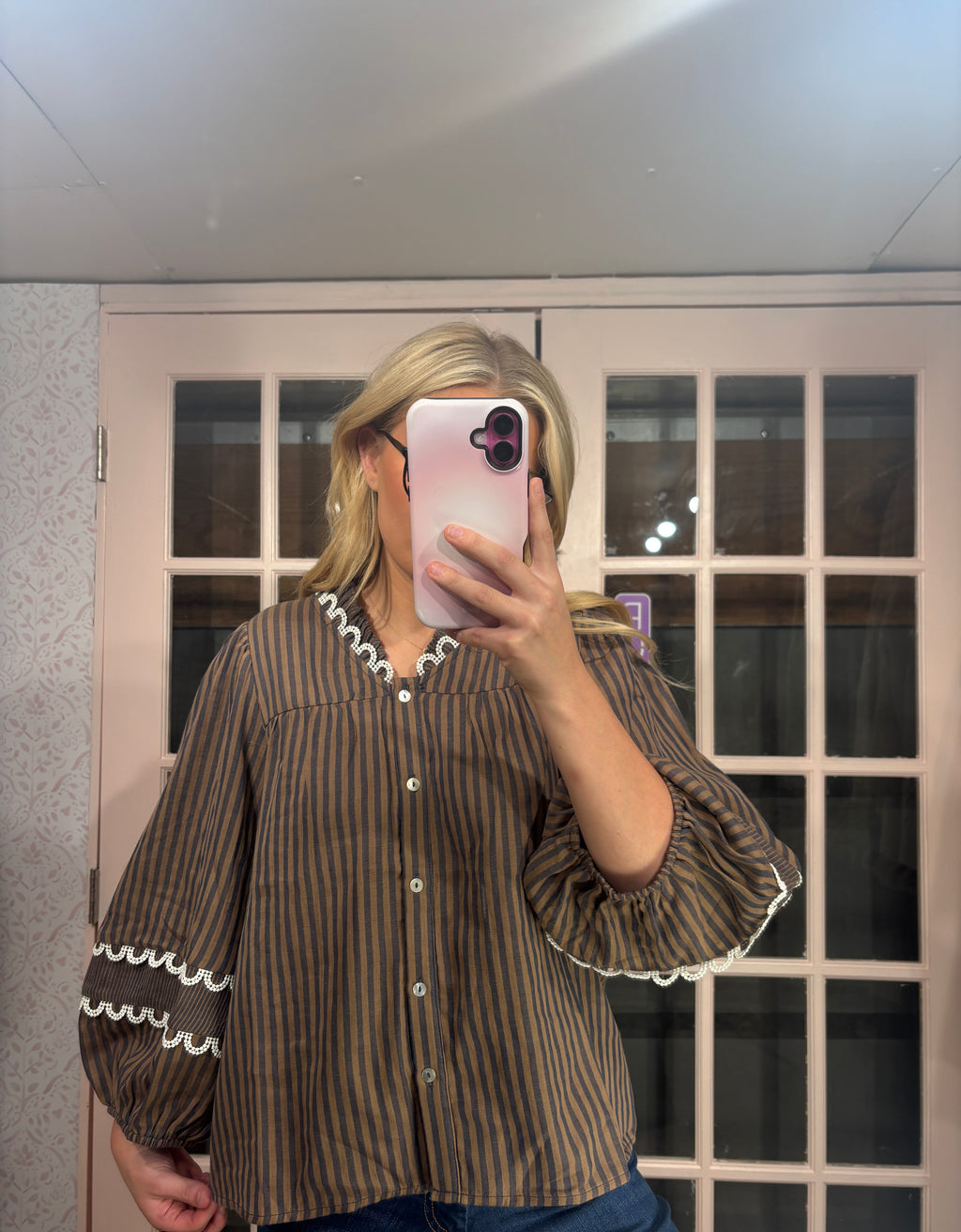 A woman takes a mirror selfie wearing a brown and black vertically striped blouse with white ruffled trim on the collar and cuffs, paired with blue jeans.