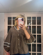A woman takes a mirror selfie wearing a brown and black vertically striped blouse with white ruffled trim on the collar and cuffs, paired with blue jeans.