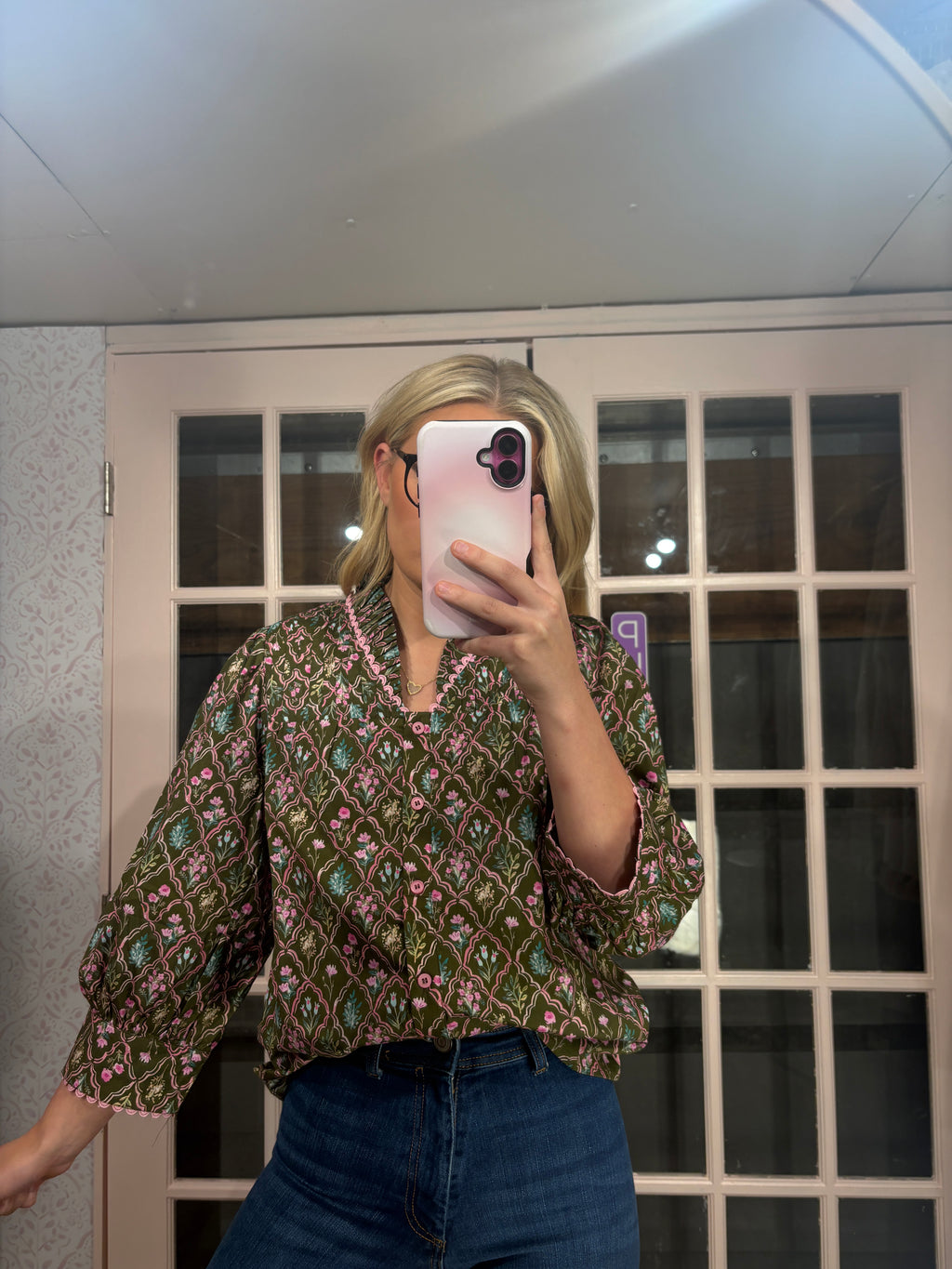 A woman takes a mirror selfie wearing a green and pink patterned button-up blouse with three-quarter sleeves and dark blue jeans, standing in front of a glass-paneled door.