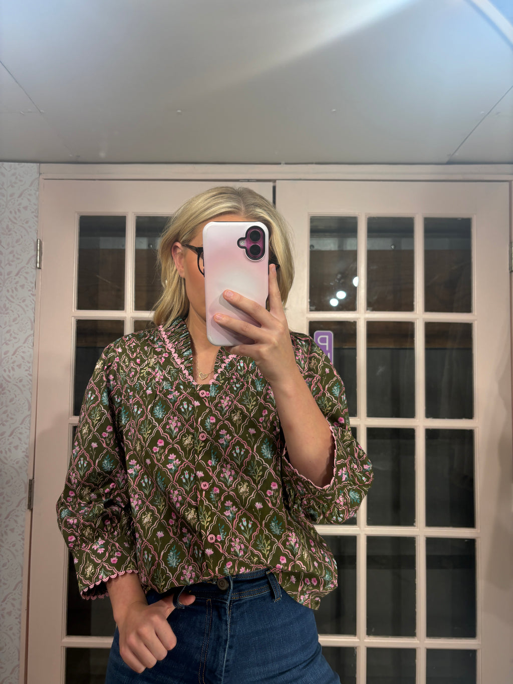 A woman takes a mirror selfie wearing a green and pink patterned blouse with blue jeans, standing in front of a white-framed glass door.