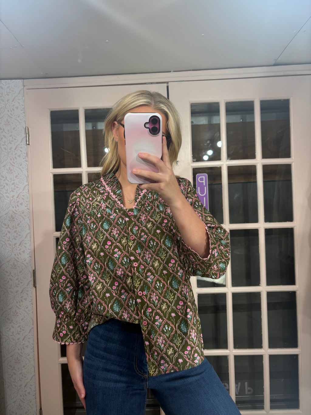 A woman takes a mirror selfie wearing a bohemian-style patterned blouse with blue jeans. The blouse features a green base with pink, orange, and blue geometric patterns.