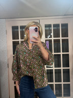 A woman takes a mirror selfie wearing a bohemian-style patterned blouse with blue jeans. The blouse features a green base with pink, orange, and blue geometric patterns.