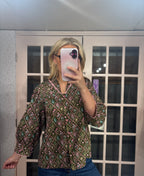 A woman takes a mirror selfie wearing a long-sleeve button-up blouse with a green, pink, and brown geometric pattern, paired with blue jeans.