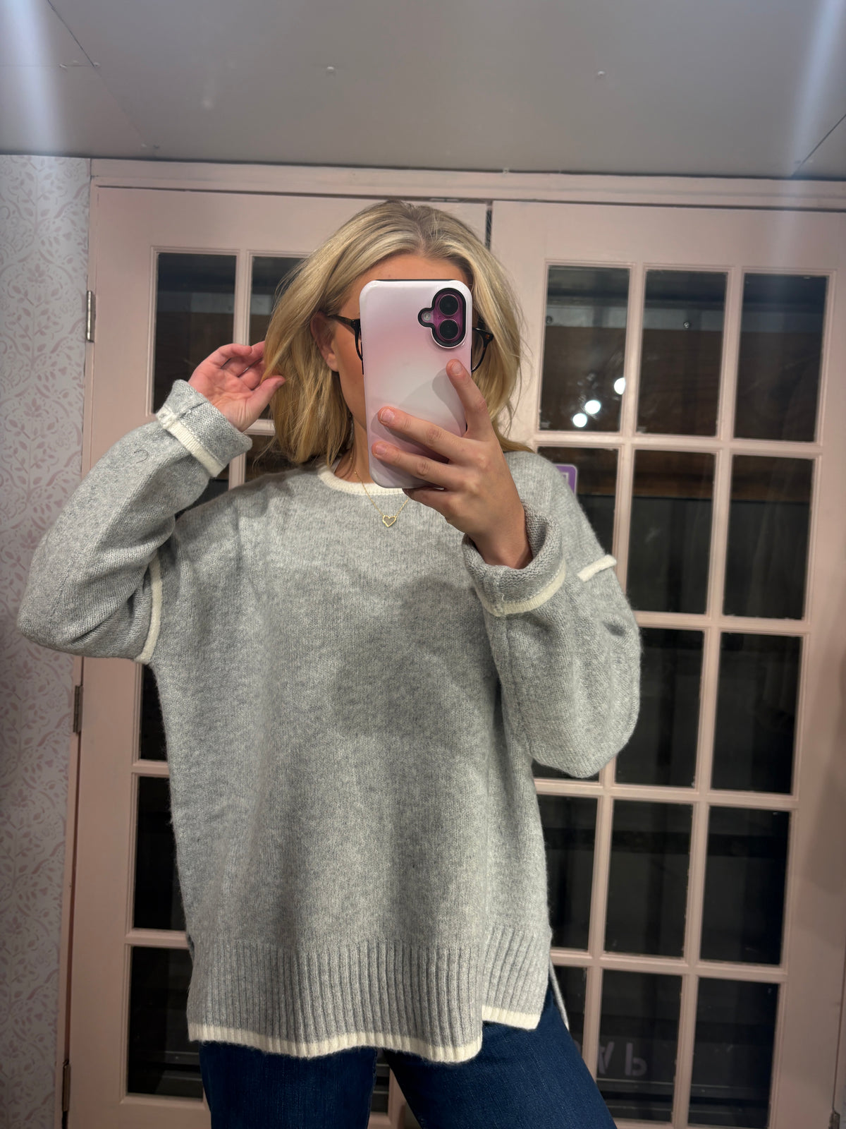 A woman takes a mirror selfie wearing a light gray knit sweater with white contrast trim, paired with dark blue jeans, in front of a glass-paneled door.