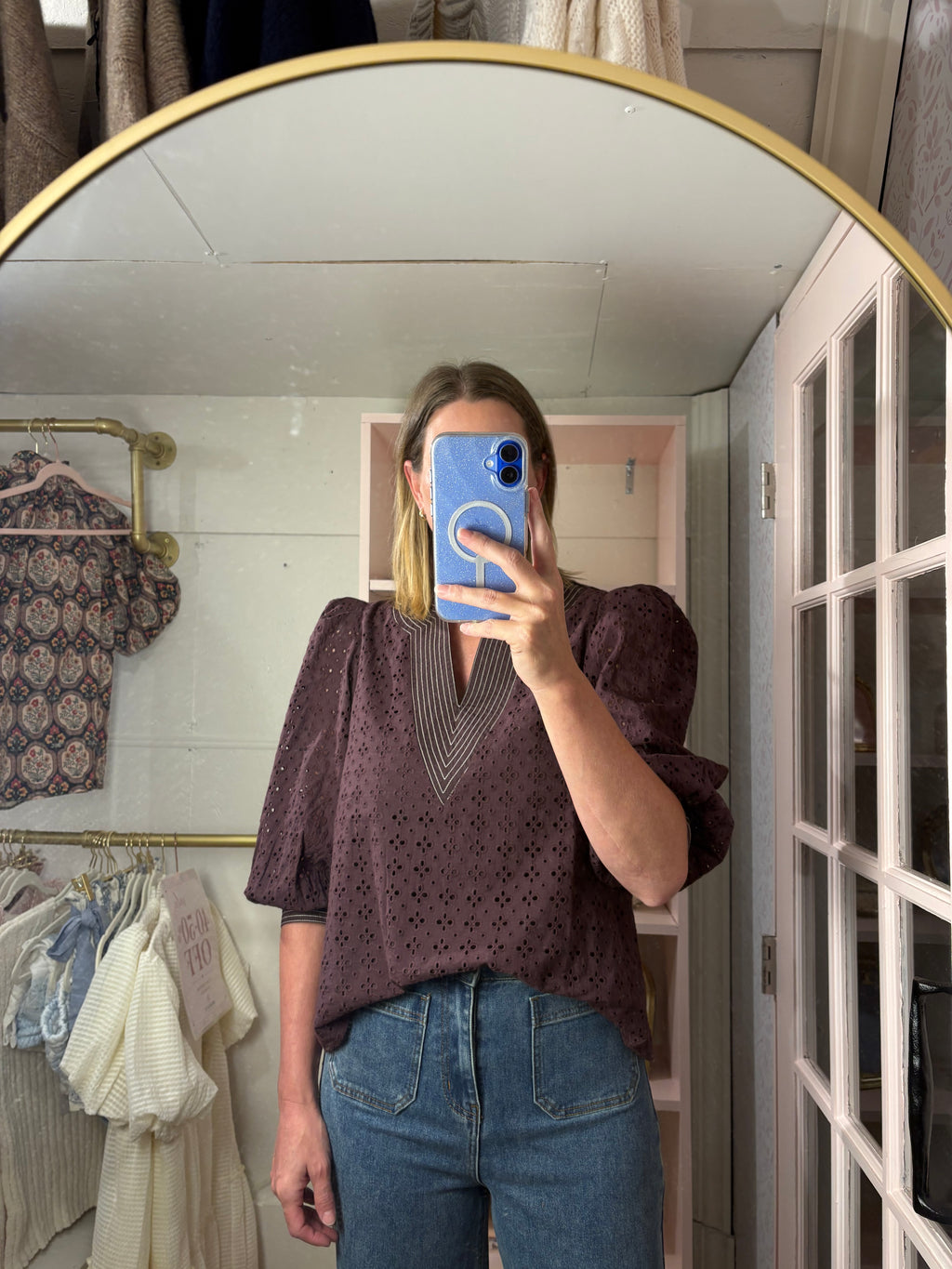 A woman takes a mirror selfie in a boutique showing a chocolate  eyelet puff-sleeve blouse with a V-neckline, paired with blue jeans.