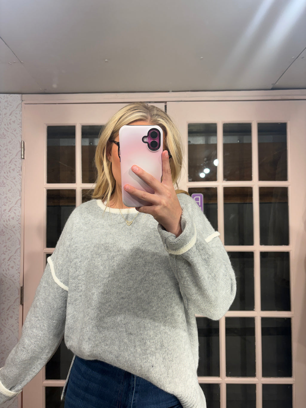 A woman takes a mirror selfie wearing a light gray knit sweater with white contrast trim and dark blue jeans, standing in front of a pink-framed glass door.